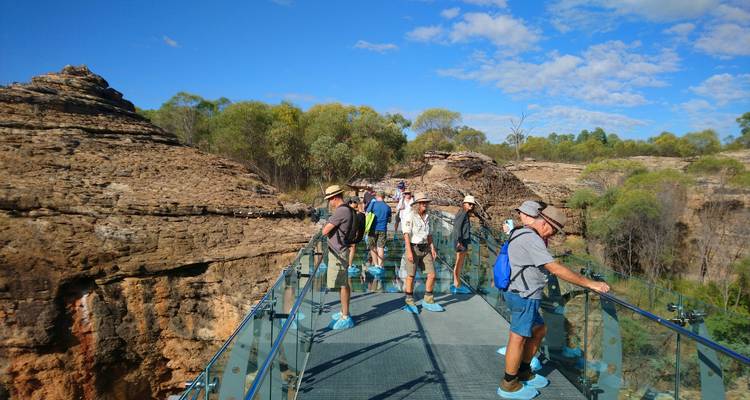 Des touristes portant des sur-chaussures se tiennent sur une passerelle de verre s'étendant au-dessus d'une gorge accidentée sous un ciel bleu éclatant.