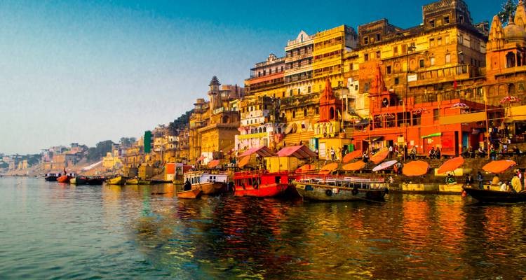 Colourful ghats and temples lining the Ganges River with boats and vivid reflections at sunrise.
