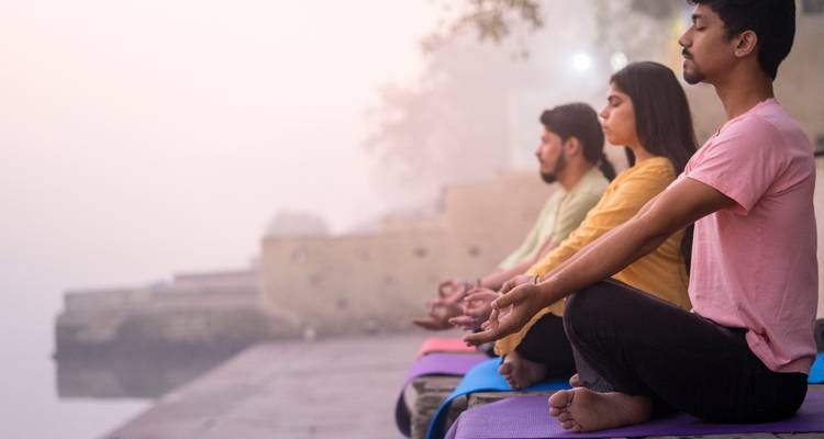 Three people practicing seated meditation on mats beside a misty riverside at dawn.