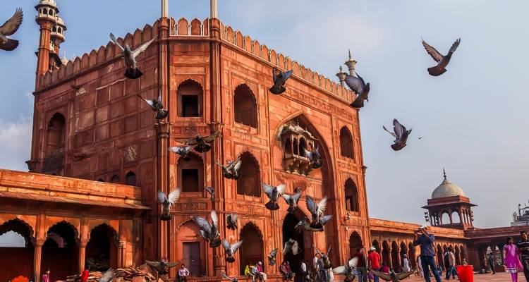Historic red-sandstone mosque with pigeons in flight against a blue sky.