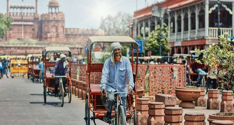 Elderly man with orange beard pedalling a cycle rickshaw past red fortifications and bustling street.