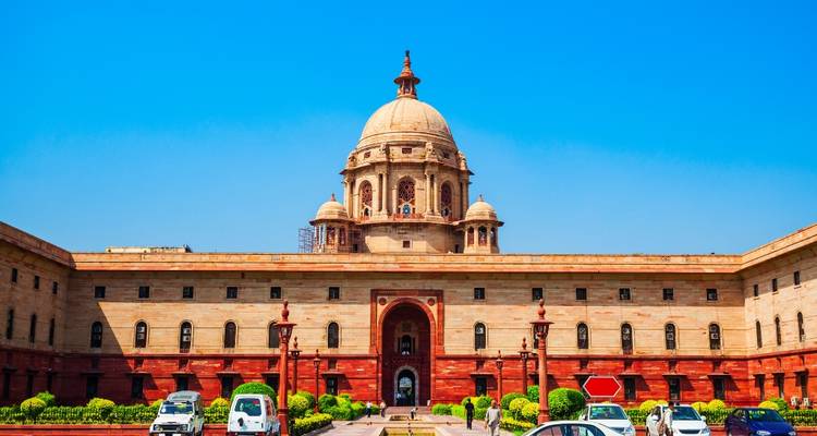 Symmetrical government building with domed roof framed by bright blue sky and parked cars in foreground.