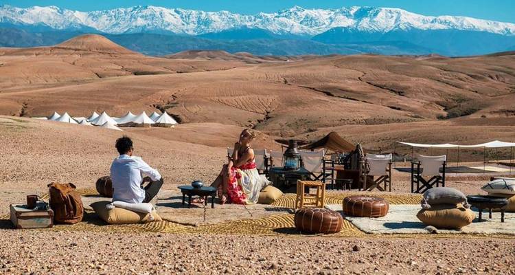 Un couple se détend dans un élégant camp désertique avec des poufs et des tapis, avec en arrière-plan des montagnes enneigées au loin sous un ciel bleu dégagé.