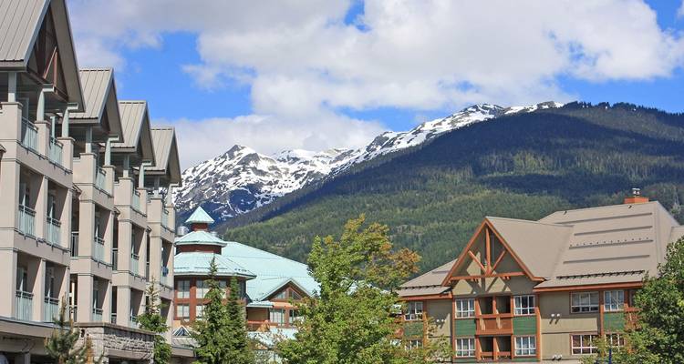 Bâtiments pittoresques du village alpin de Whistler avec chaîne de montagnes enneigées en arrière-plan sous un ciel partiellement nuageux.