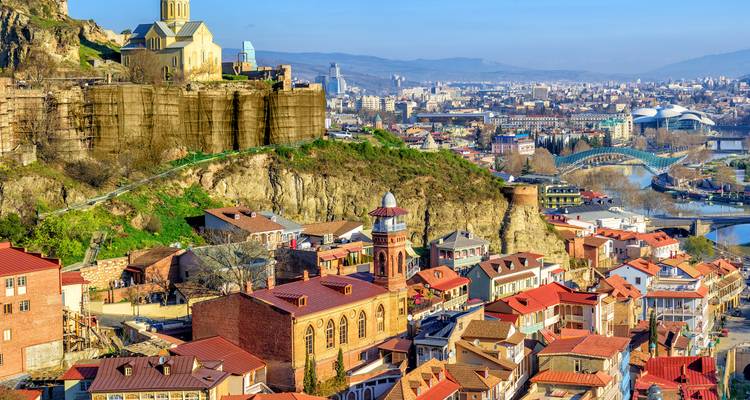 Farbenfrohe Dächer der Altstadt von Tiflis reihen sich unterhalb einer Klippenkirche und Festung, mit moderner Stadtlandschaft dahinter.