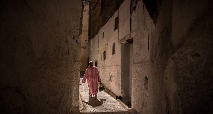 Woman in traditional dress walking through a dim, narrow stone alleyway lit by a shaft of sunlight