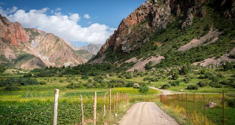Dirt road winds through a lush green valley surrounded by rugged red and grey mountains under blue sky.