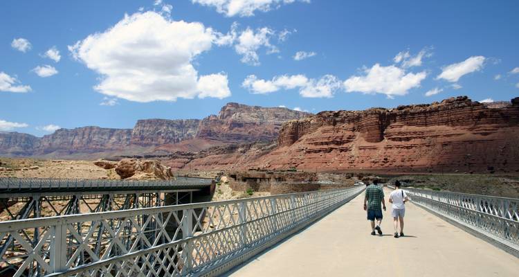 Two travelers walk along a metal truss bridge spanning a deep desert canyon with red rock cliffs.