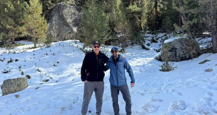 Two men smile while standing on snow-covered ground among tall pine trees.