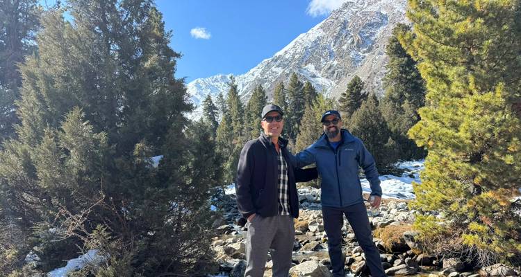 Two men pose beside a rocky creek surrounded by conifer trees and distant snowy peaks.
