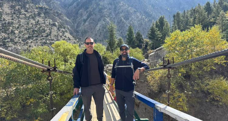 Two travelers stand on a suspension bridge with rocky mountains and autumn foliage behind.