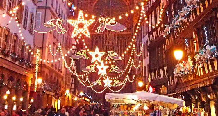 Warm strings of festive lights and star motifs illuminate a bustling Strasbourg Christmas market street at dusk.
