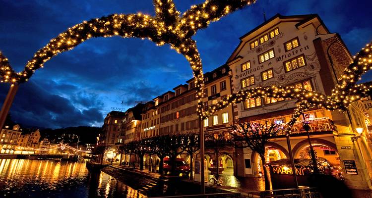 Twinkling holiday lights arch over Lucerne’s waterfront buildings reflecting in the Reuss River at blue hour.
