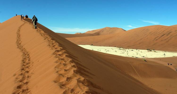 Travellers hike up a towering red sand dune with a ridgeline footpath against a vivid blue desert sky.