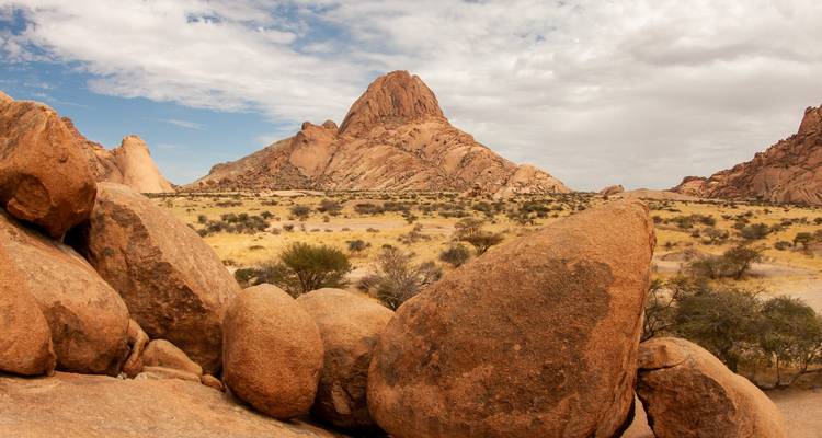 Massive orange-pink granite boulders and a sharp peak rise above a semi-arid savanna under partly cloudy skies.