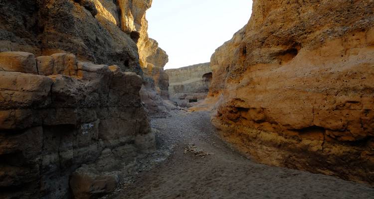 Narrow rocky canyon with sandy floor illuminated by warm sunlight filtering between its high walls.