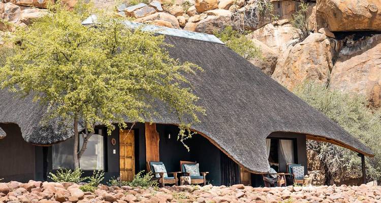 Thatched-roof safari lodge nestled among boulders and acacia trees in a semi-arid landscape.