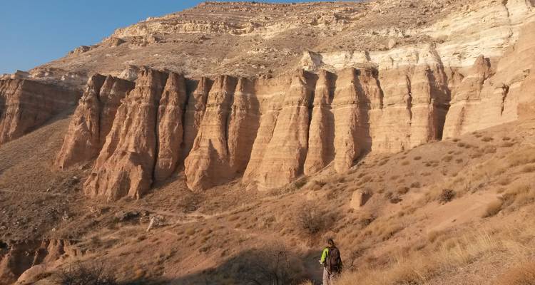 Un randonneur solitaire marche sous des falaises rocheuses stratifiées imposantes dans un canyon poussiéreux.