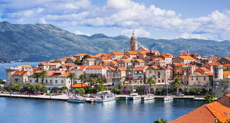 Charming medieval town with terracotta rooftops and a central bell tower set against green mountains and a blue bay.
