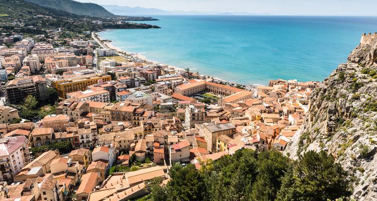 Panoramic view over the seaside town of Cefalù with terracotta roofs and turquoise coastline framed by cliffs.