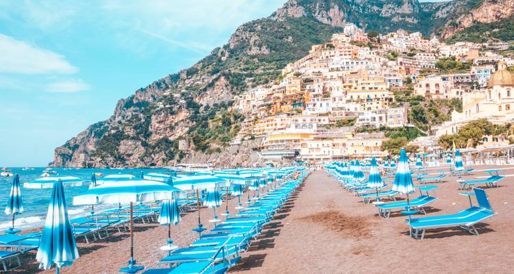 Lange Reihe blauer Sonnenschirme und Liegestühle am Strand von Positano mit dem bunten Klippendorf, das dahinter aufsteigt.
