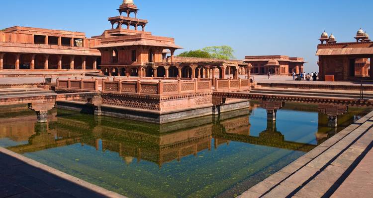 Les pavillons en grès rouge de Fatehpur Sikri se reflètent dans un bassin rectangulaire sous un ciel dégagé.
