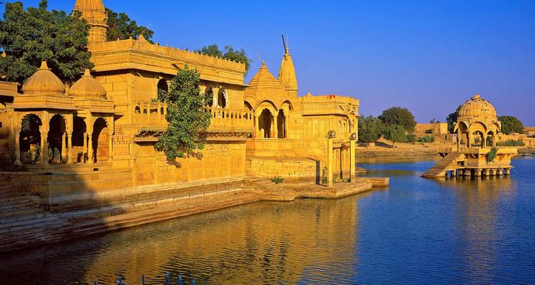 Pittoreske Gadisar Lake tempels in Jaisalmer die gouden gloeien naast kalm water onder een blauwe hemel.