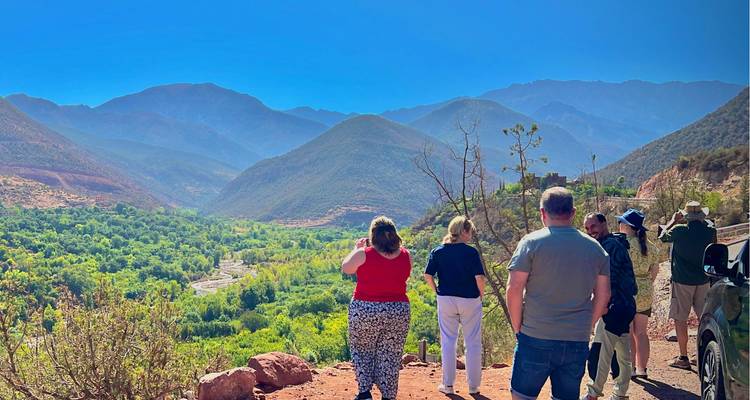Tourists standing at a roadside viewpoint overlooking a verdant valley backed by rugged Atlas Mountains