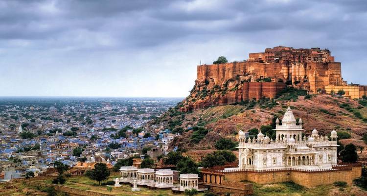 Dramatic view of Mehrangarh Fort atop a rusty cliff with white marble Jaswant Thada and blue city sprawling below