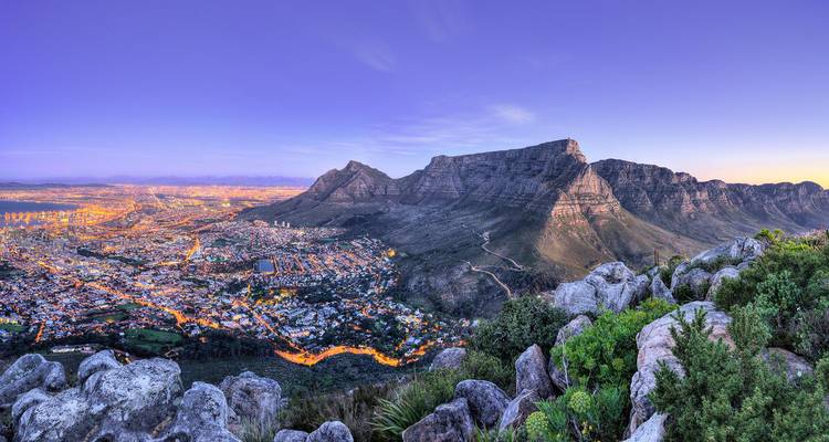 Sweeping twilight panorama of Cape Town’s illuminated city bowl beneath flat-topped Table Mountain viewed from a rocky lookout.