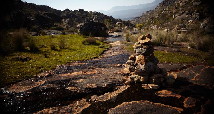 Rock cairn beside a small stream flowing through rugged Cederberg mountains under hazy light.