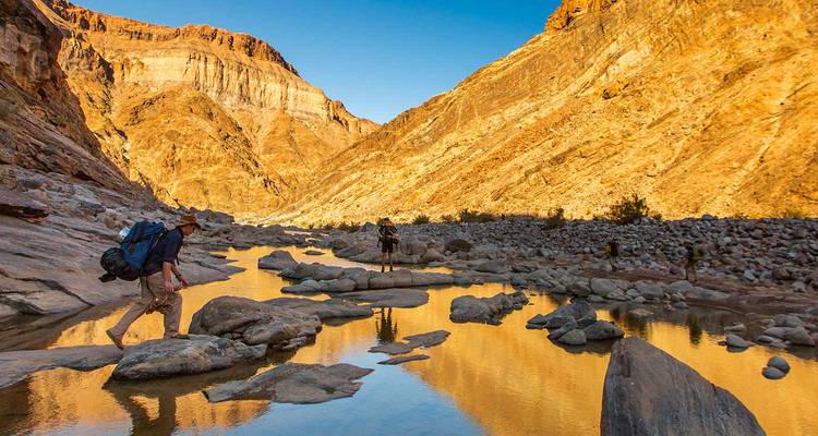 Backpackers negotiate stepping stones along the reflective river floor of Fish River Canyon’s golden walls.