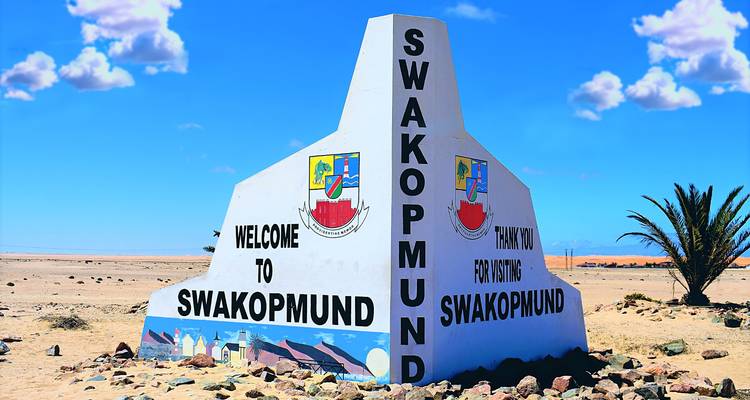 Large white concrete roadside monument welcoming and thanking visitors to Swakopmund in the desert.