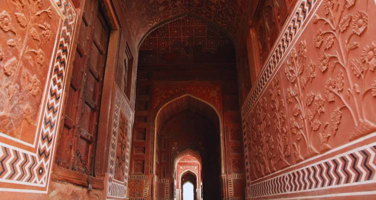 Red sandstone corridor richly carved with floral and zig-zag patterns leading to distant arches.