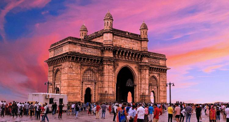 The Gateway of India monument stands against a vivid pink-purple sunset sky as crowds gather around its arch.