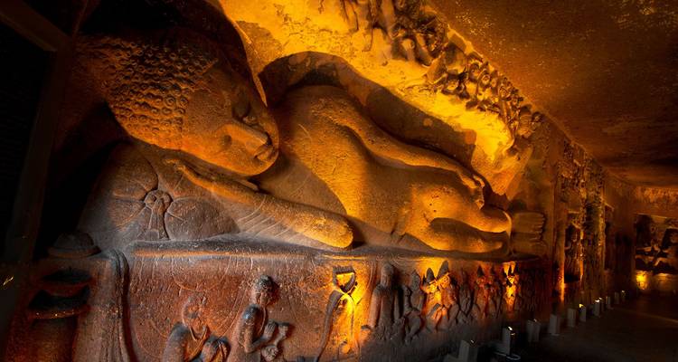 A softly lit reclining Buddha carved into rock exudes serenity inside a dimly illuminated cave hall.