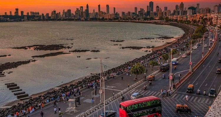 Mumbai’s seafront skyline glows orange at sunset as traffic moves along Marine Drive and crowds stroll the promenade.