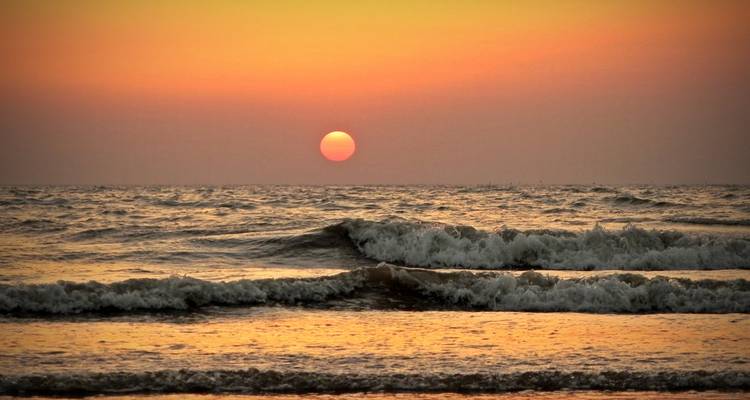 A round orange sun sinks toward gentle ocean waves creating a tranquil seascape at dusk.