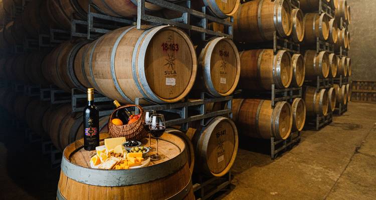 Wine barrels stack high in a cellar where a barrel top displays cheese, fruit and a glass of red wine.