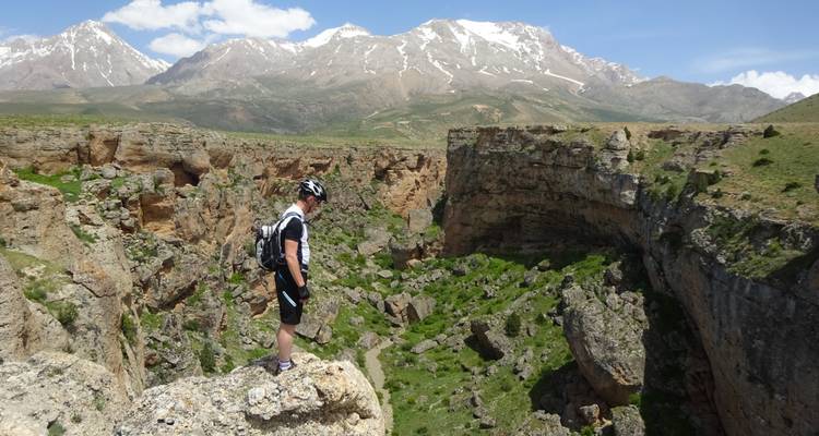 Cycliste debout au bord d'une falaise rocheuse surplombant un canyon spectaculaire et des sommets enneigés dans les Aladağlar.