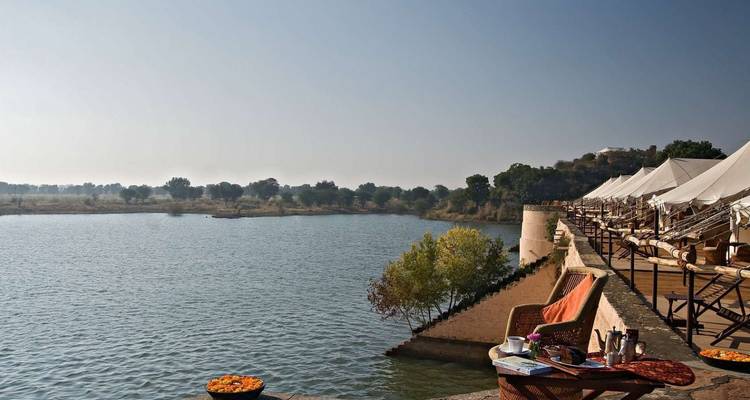 Luxury lakeside tented camp with deck chairs beside calm reservoir in rural Rajasthan.