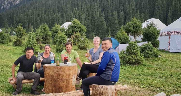 Travellers relax with drinks on rustic log stools outside yurts in a forested mountain camp.