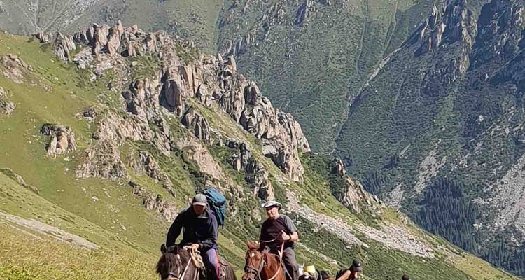 Horseback riders ascend a steep grassy slope with rocky cliffs towering above.