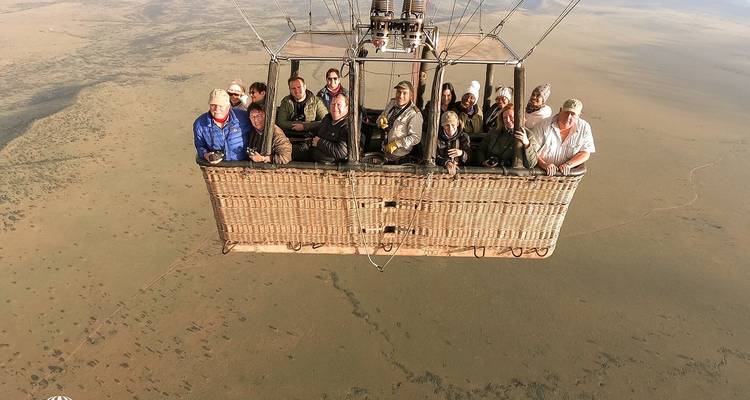 A full basket of safari tourists smiles for the camera while the balloon drifts over endless African plains.