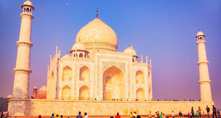 Taj Mahal glowing in warm evening light against a pastel sky, crowds gathered at its base.