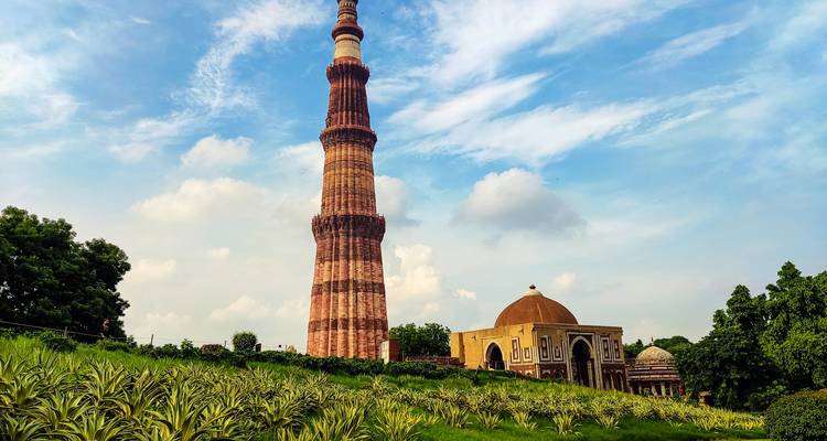 Soaring Qutub Minar tower framed by green gardens under a bright blue sky with puffy clouds.