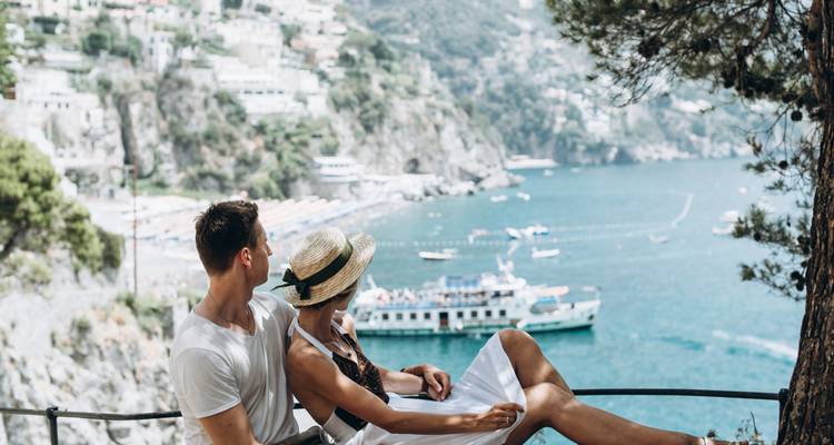 Couple se relaxant sur un banc avec vue sur un littoral turquoise parsemé de bateaux et de falaises spectaculaires.