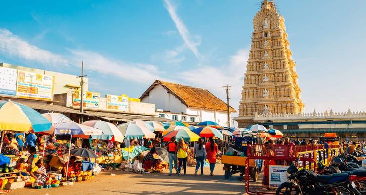 Marché de rue animé avec des parasols colorés à côté d'une tour gopuram ornée sous un ciel bleu.