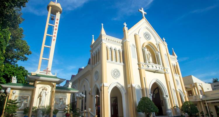 Yellow-cream colonial cathedral with tall bell tower gleams under clear blue Vietnamese sky.