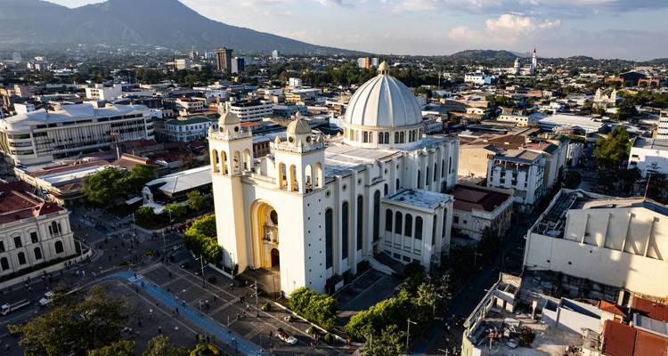 Aerial view of San Salvador city with its white domed Metropolitan Cathedral dominating the skyline and a distant volcano.